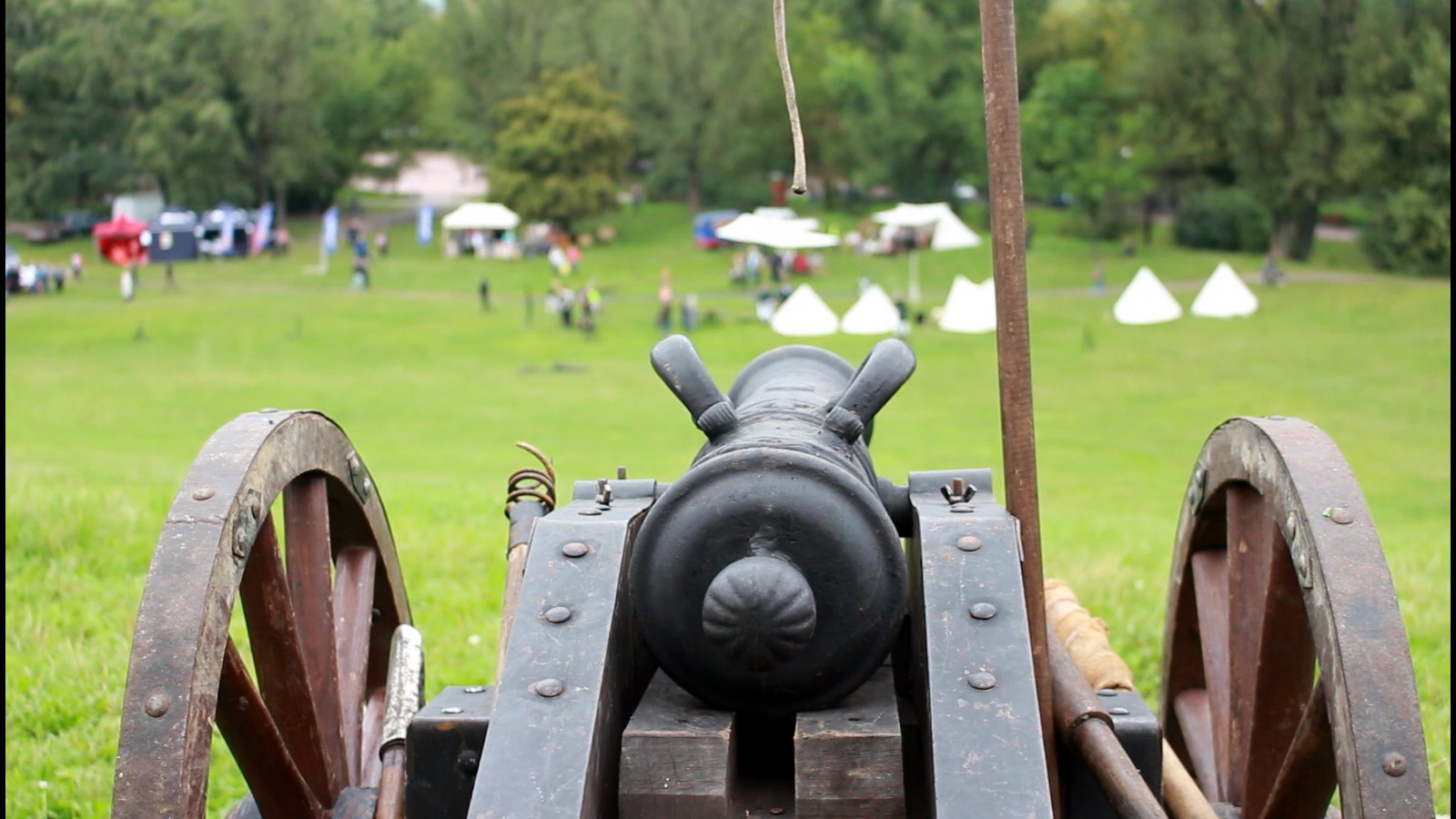 Czarna armata z drewnianymi kołami na trawiastym terenie podczas wydarzenia plenerowego z białymi namiotami i ludźmi w tle.