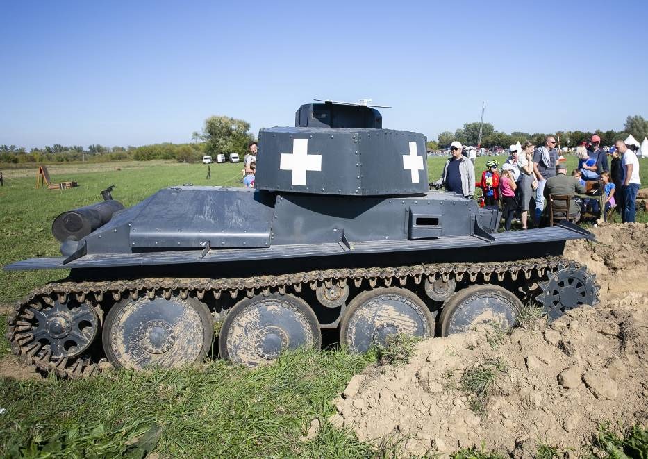 Szary, lekko zardzewiały czołg z białymi krzyżami na wieżyczce, częściowo zagrzebany w ziemi, na tle łąki i zgromadzenia ludzi.