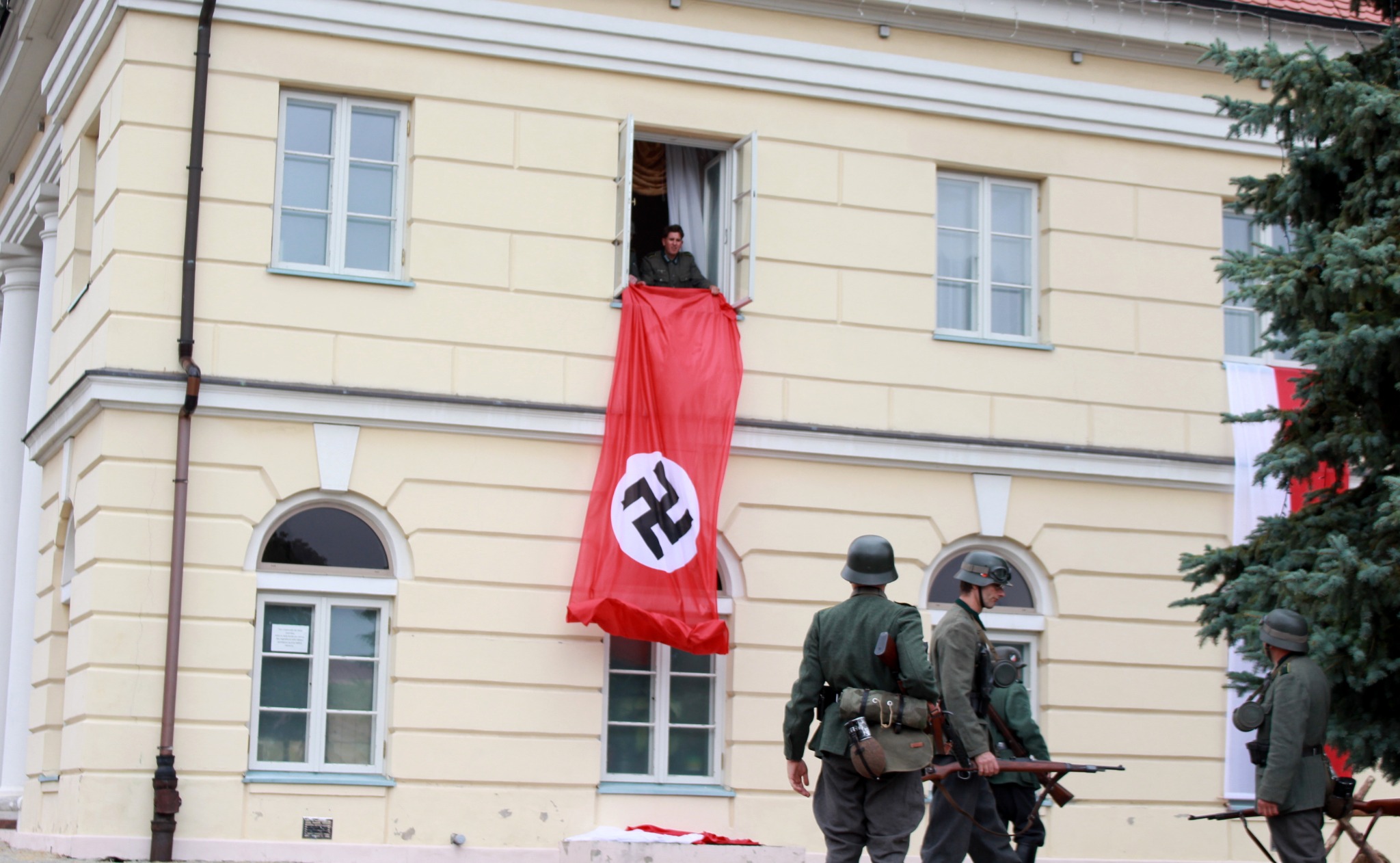 Osoba w oknie rozwiesza czerwoną flagę z symbolem na budynku, w tle żołnierze w mundurach z karabinami.