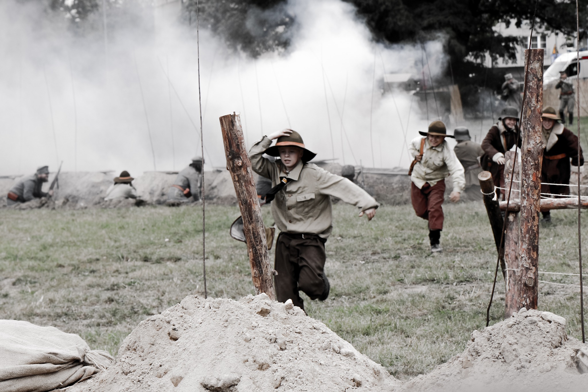 Scena rekonstrukcji historycznej bitwy z udziałem aktorów w strojach z epoki, biegających w scenerii dymu i okopów.