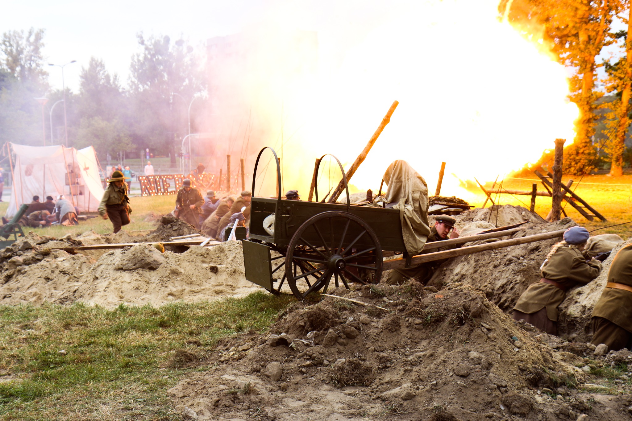 Rekonstrukcja historyczna bitwy: żołnierze w okopach, w tle wybuch i dym, wóz z osprzętem.