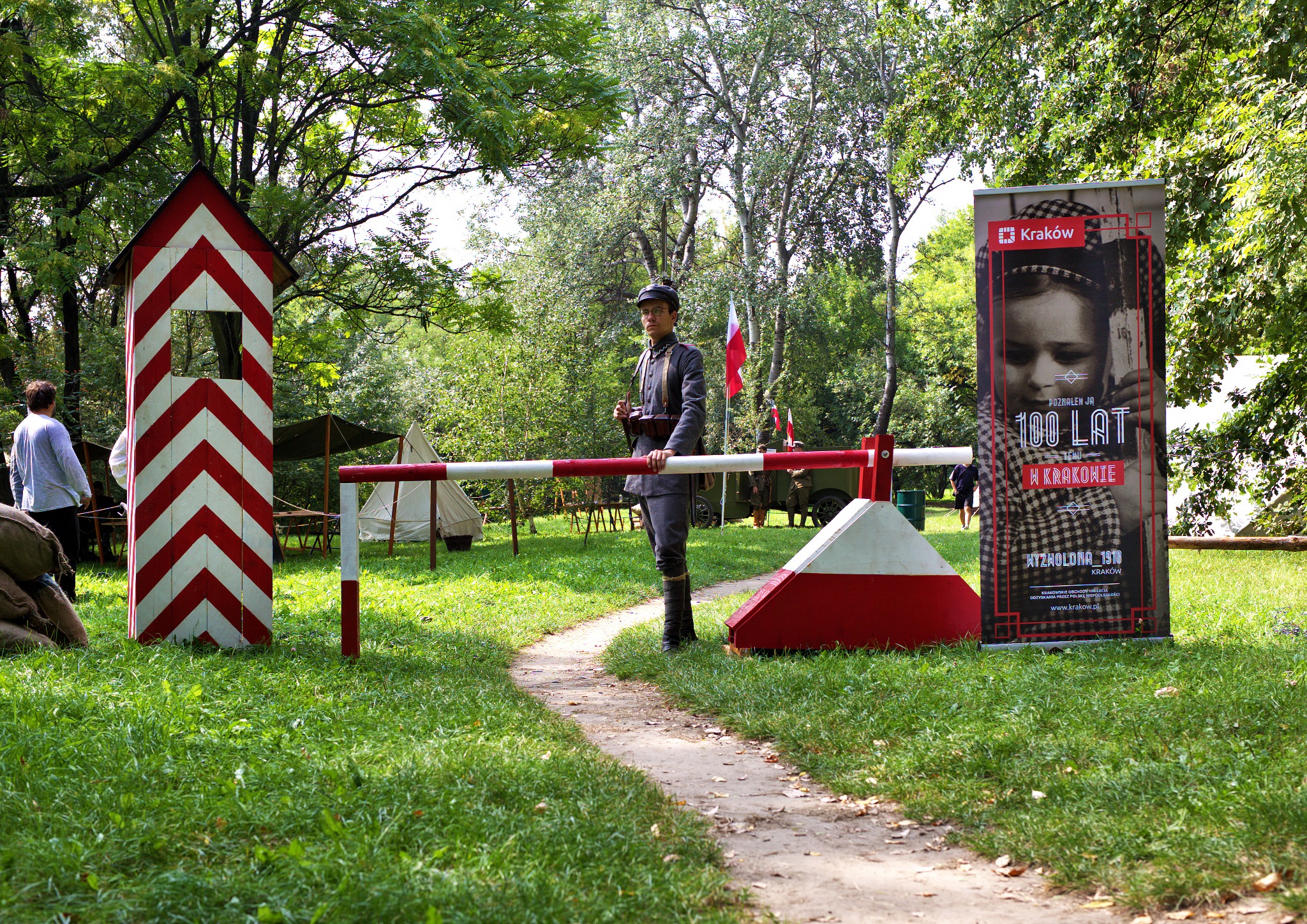 Rekonstrukcja historyczna: żołnierz w mundurze z epoki stoi przy szlabanie w biało-czerwone pasy, w tle obóz rekonstruktorów i baner reklamujący obchody 100-lecia odzyskania niepodległości w Krakowie.