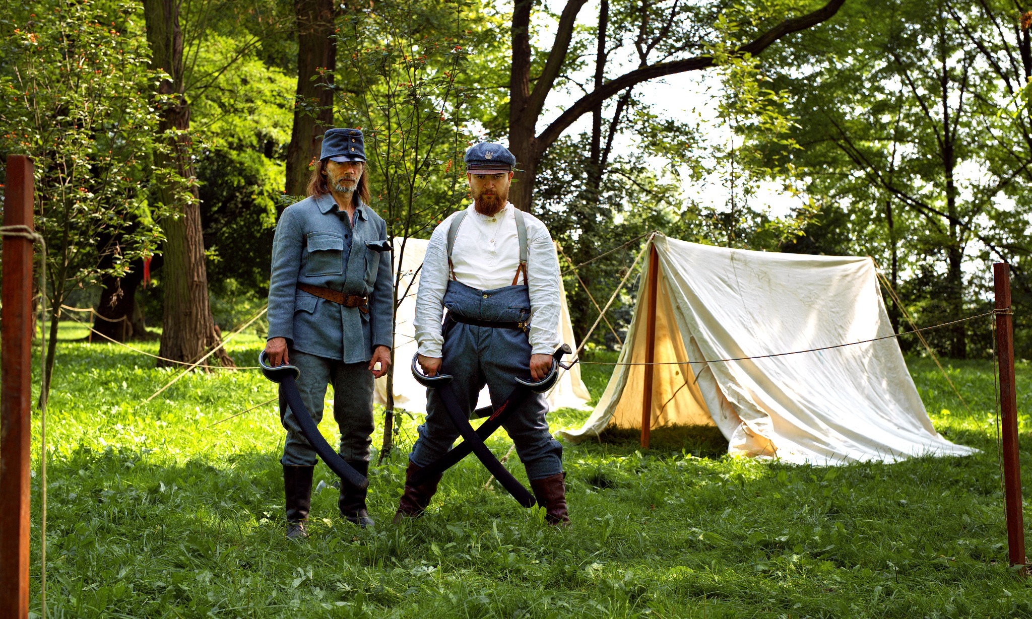 Dwóch mężczyzn w historycznych mundurach z szablami na tle namiotu w plenerze.
