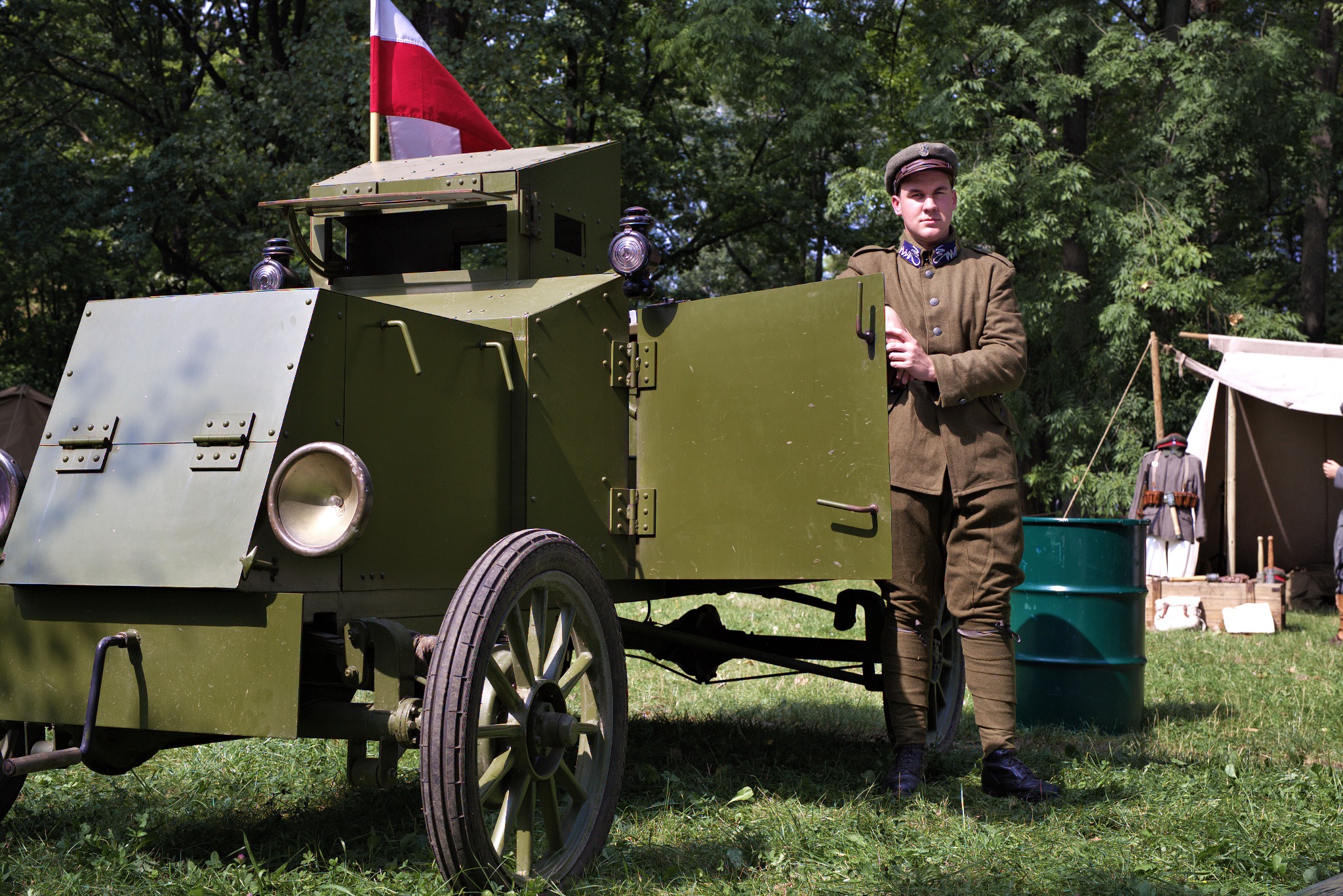 Replika polskiego samochodu pancernego z okresu I wojny światowej z żołnierzem w mundurze z epoki, flaga Polski na dachu, obóz wojskowy w tle.