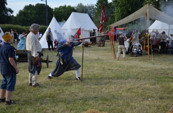 Mężczyzna w stroju historycznym strzela z muszkietu podczas rekonstrukcji bitwy na trawiastym terenie, w tle białe namioty i widzowie.