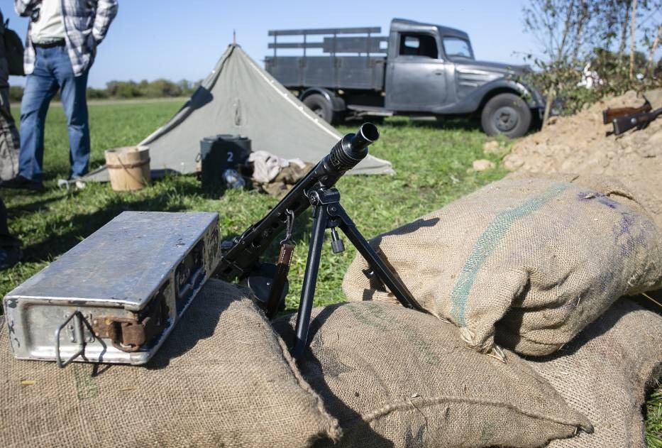 Rekonstrukcja historyczna: karabin maszynowy na workach z piaskiem, wojskowy namiot i ciężarówka w tle, osoba w cywilnym ubraniu w lewym górnym rogu.