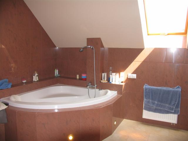 Wykończona łazienka na poddaszu z narożną wanną, brązowymi płytkami i oknem dachowym wpadającym w kadr.