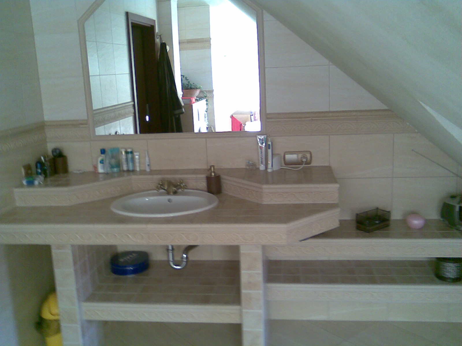 Umywalka z beżowymi płytkami w łazience na poddaszu, z widocznym lustrem, półkami i akcesoriami łazienkowymi.