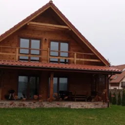 Drewniany dom z czerwonym dachem i zadaszonym tarasem, na którym stoi grill i drewniane meble. W tle widoczny mniejszy budynek o podobnej architekturze, a także inny dom z białymi ścianami.