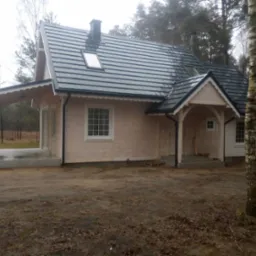 Jasny, parterowy dom z ciemnoszarą dachówką, widoczny z zewnątrz, z elementami wykończeniowymi w kolorze białym. Do domu przylega zadaszony taras z betonową wylewką. W tle widoczne drzewa.