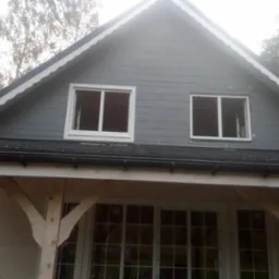 Częściowo wykończony, dwukondygnacyjny budynek z szarego drewna z czarnym dachem i białymi ramami okiennymi. Widoczne niezabudowane otwory okienne na poddaszu i drewniane wsporniki werandy.