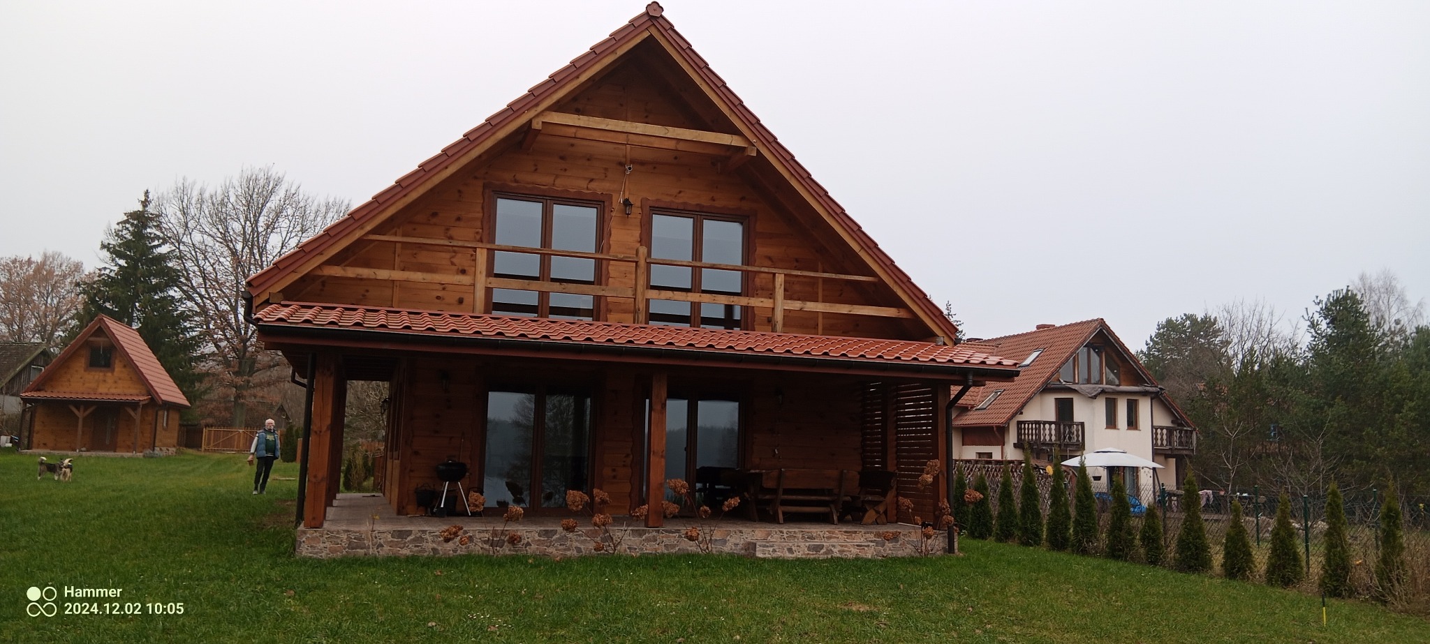 Drewniany dom z czerwonym dachem i zadaszonym tarasem, na którym stoi grill i drewniane meble. W tle widoczny mniejszy budynek o podobnej architekturze, a także inny dom z białymi ścianami.