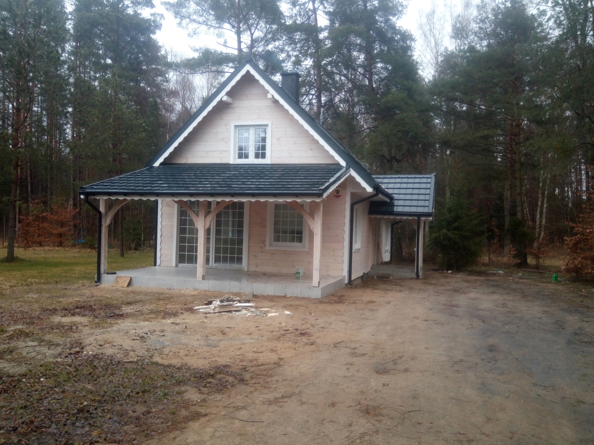 Jasny dom szkieletowy z ciemnym dachem, werandą i widoczną więźbą dachową, usytuowany na działce z drzewami iglastymi w tle, częściowo wykończony teren wokół domu.