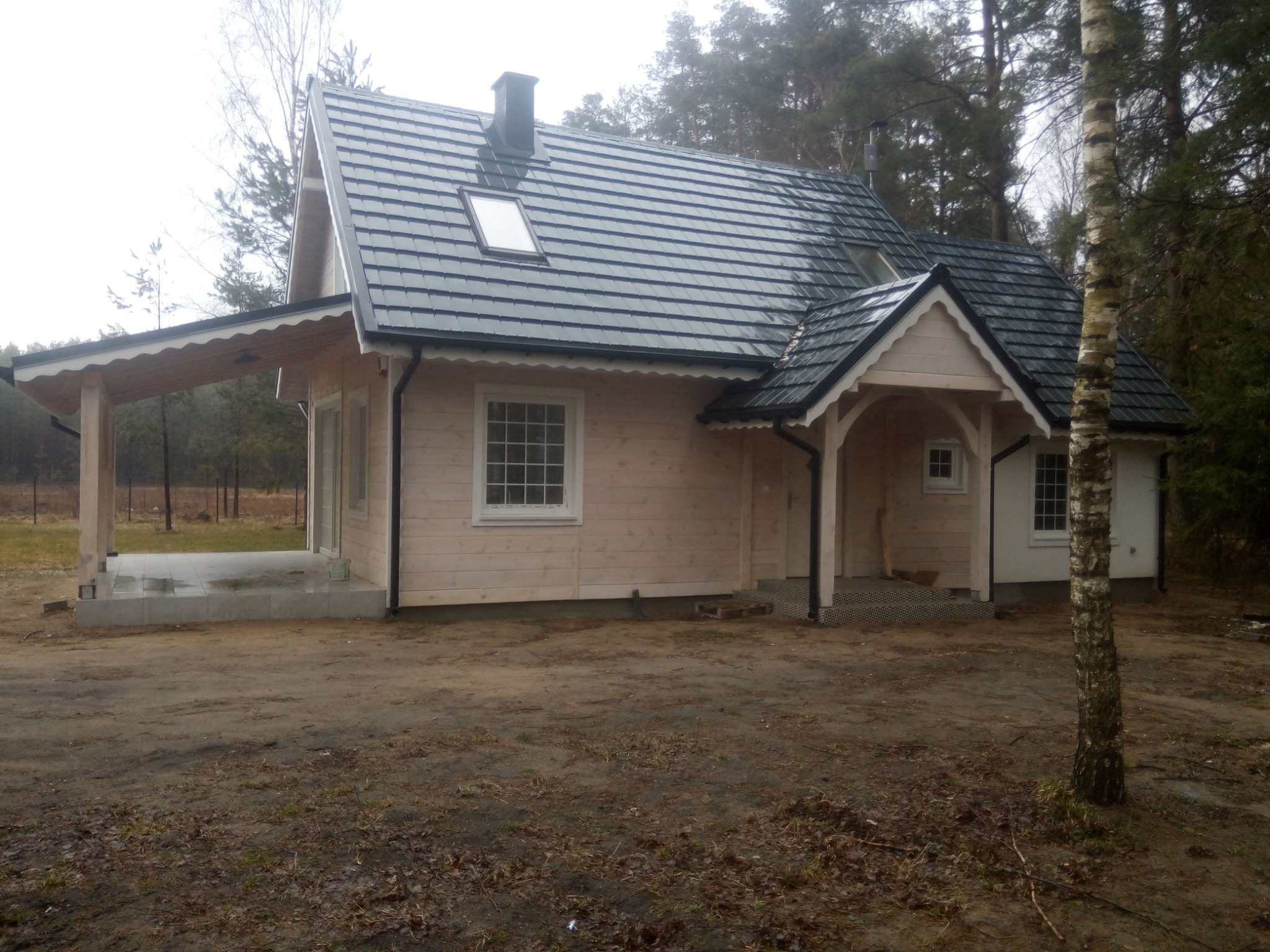 Jasny, parterowy dom z ciemnoszarą dachówką, widoczny z zewnątrz, z elementami wykończeniowymi w kolorze białym. Do domu przylega zadaszony taras z betonową wylewką. W tle widoczne drzewa.