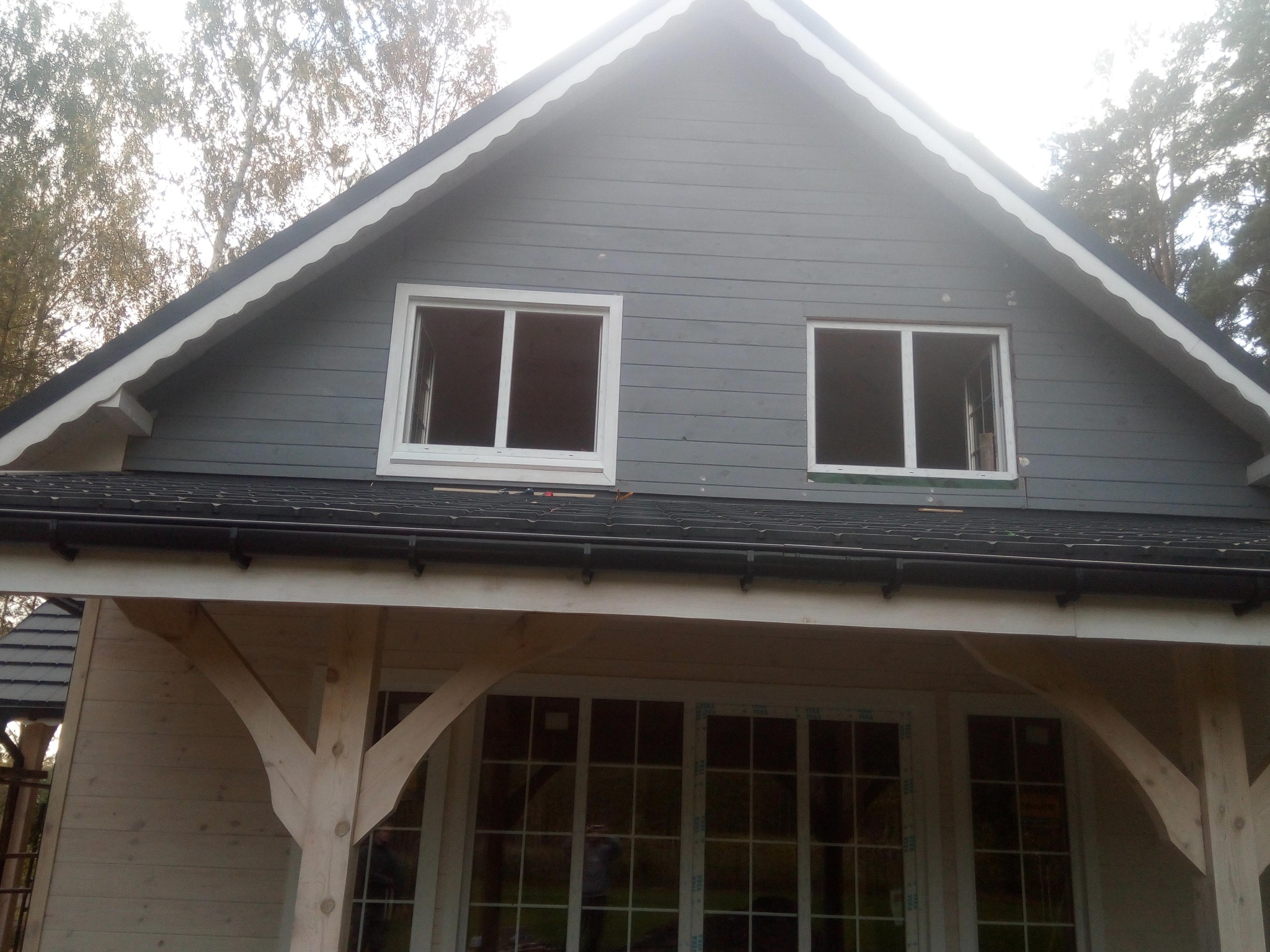 Częściowo wykończony, dwukondygnacyjny budynek z szarego drewna z czarnym dachem i białymi ramami okiennymi. Widoczne niezabudowane otwory okienne na poddaszu i drewniane wsporniki werandy.