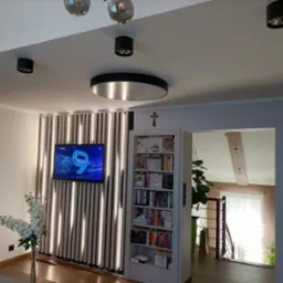 Salon z telewizorem na ścianie z pionowych paneli oświetleniowych, regałem na książki z krzyżykiem na górze i widokiem na schody w tle.