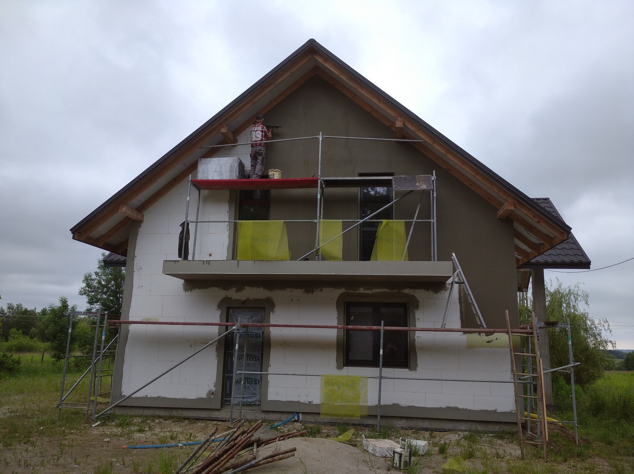 Dom w Kolbuszowej w trakcie prac elewacyjnych: ocieplanie i nakładanie tynku, widoczne rusztowania i pracownik zacierający ścianę na wysokości poddasza.