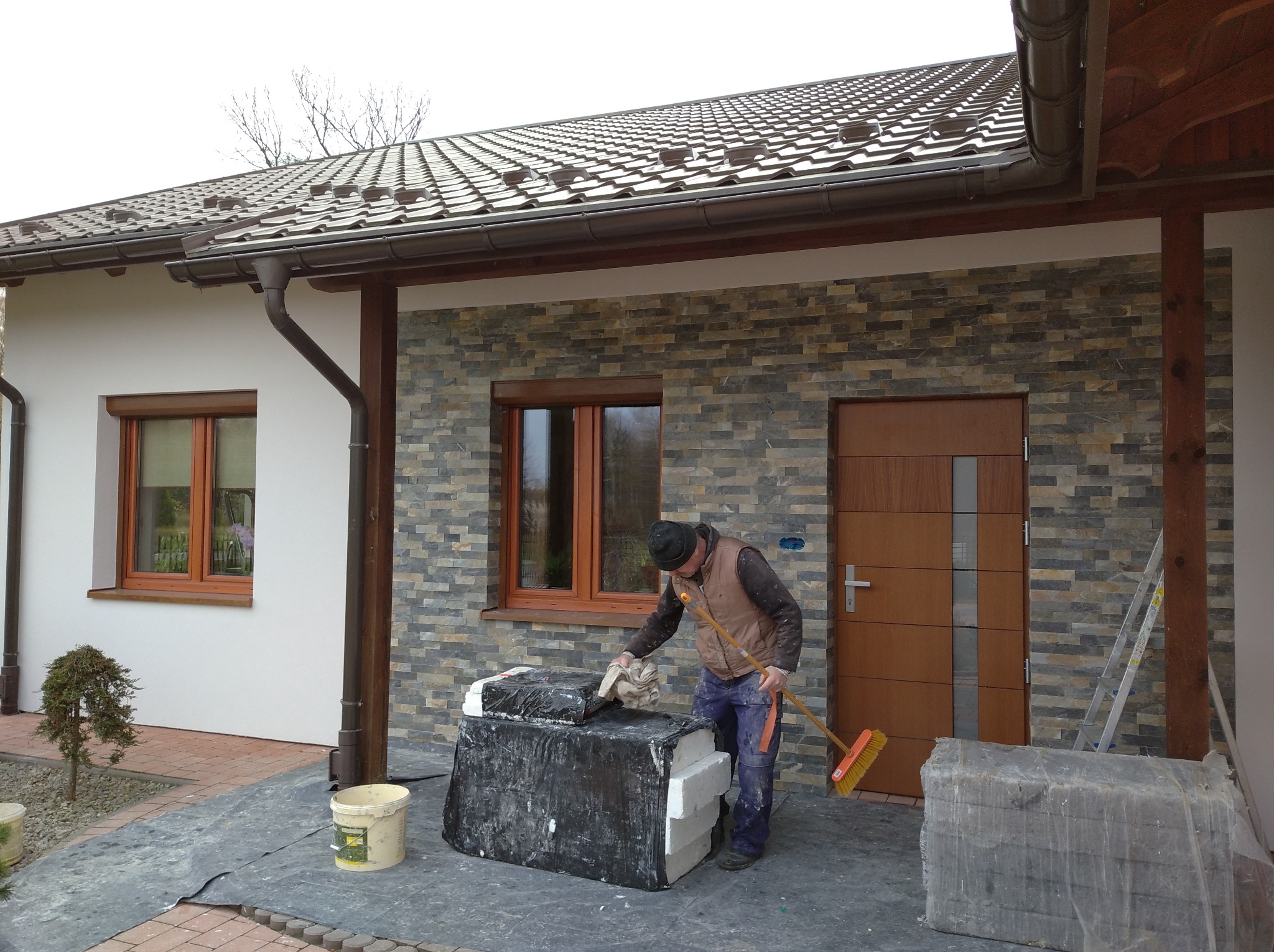 Pracownik w kamizelce i czapce czyści powierzchnię palety z materiałami budowlanymi przed budynkiem z elewacją z kamienia, widoczne okno i drzwi wejściowe.