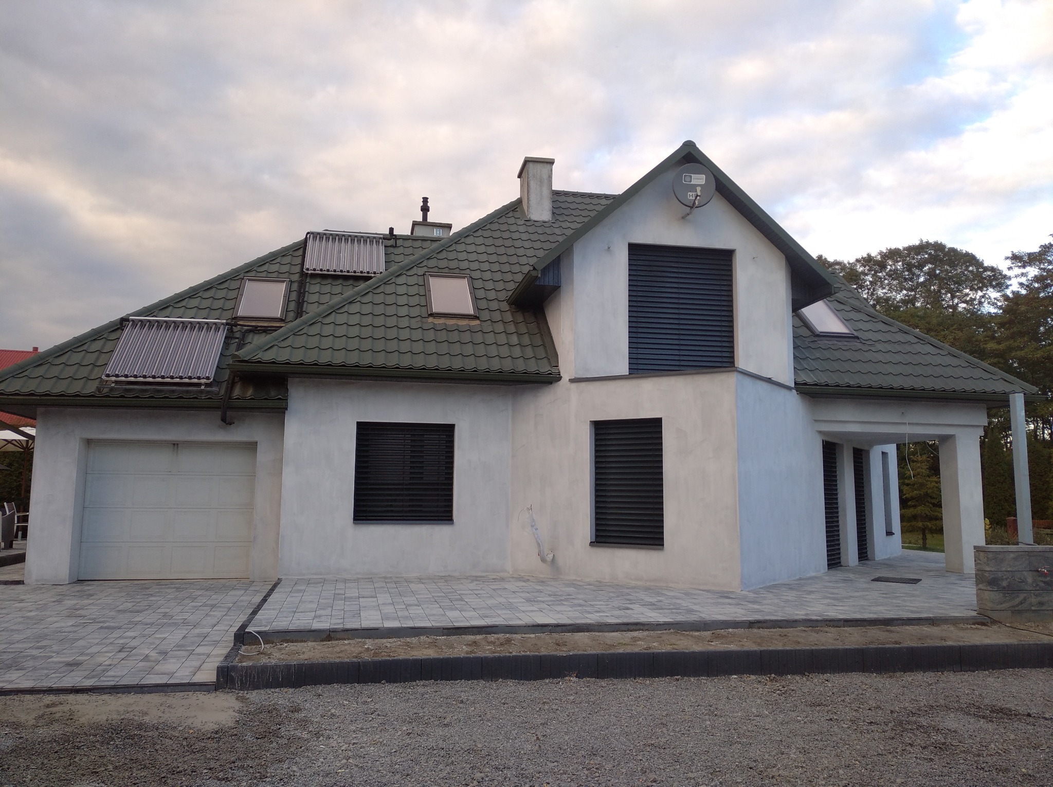 Nowoczesny dom jednorodzinny z szarą elewacją, ciemnozielonym dachem z kolektorami słonecznymi i kostką brukową na podjeździe, sfotografowany w pochmurny dzień.