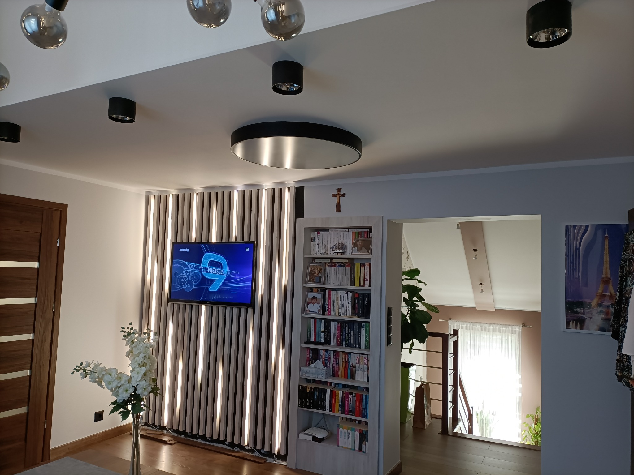 Salon z telewizorem na ścianie z pionowych paneli oświetleniowych, regałem na książki z krzyżykiem na górze i widokiem na schody w tle.