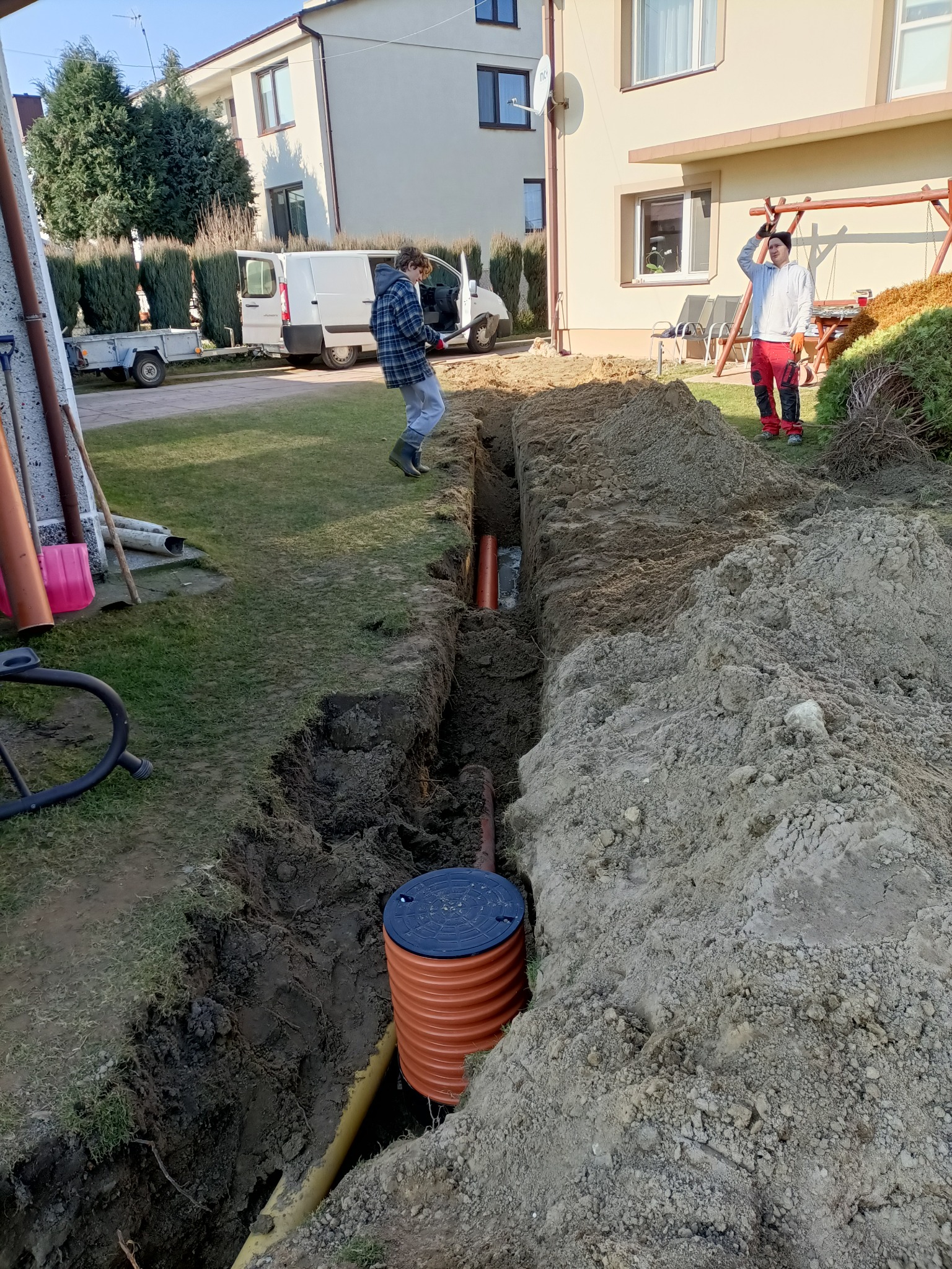 Wykop pod instalację kanalizacyjną na trawniku przed domem, widoczne rury, studzienka z pokrywą i dwie osoby.