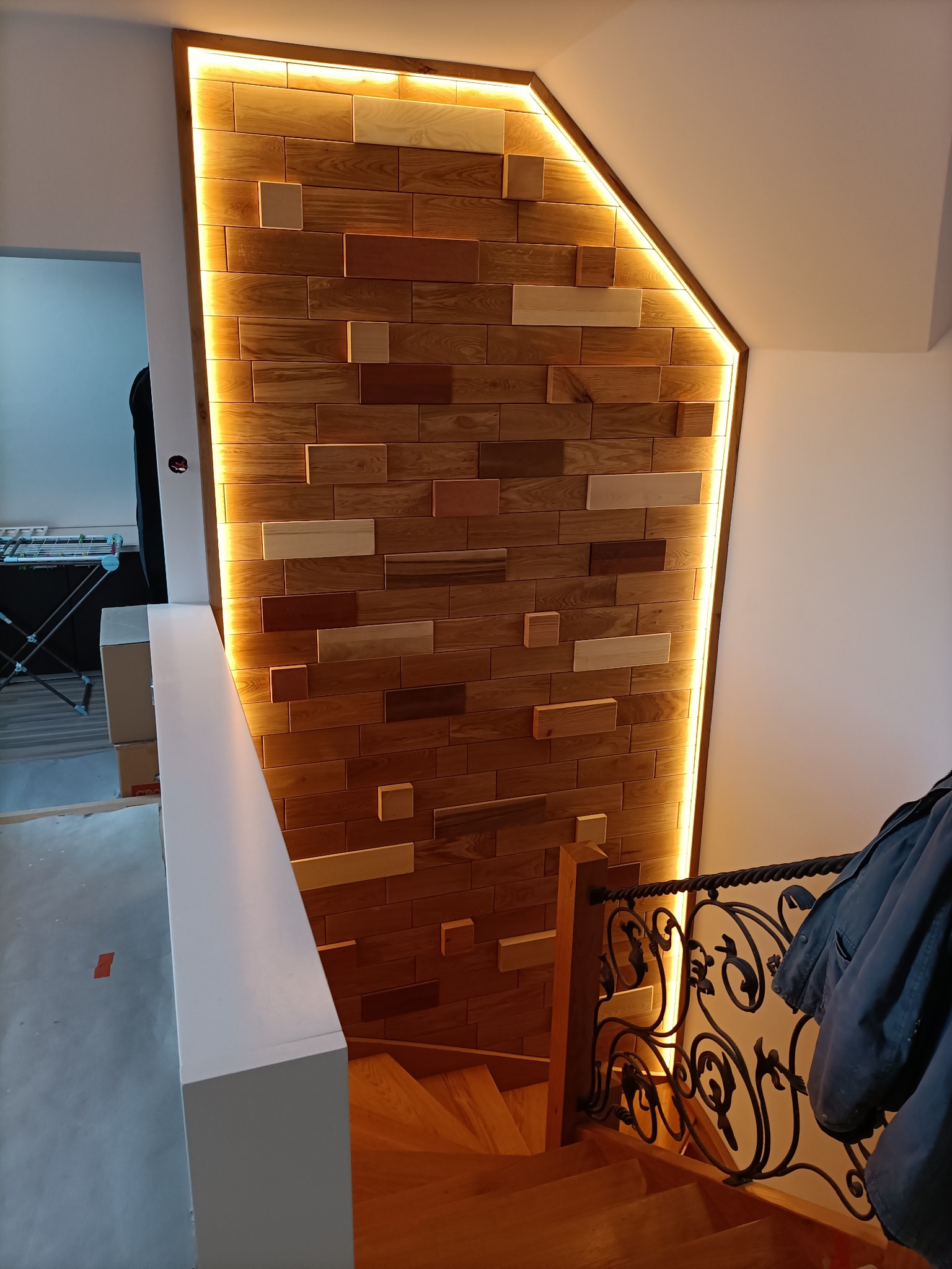 Ściana dekoracyjna z drewnianych, prostokątnych elementów o różnej barwie, podświetlona taśmą LED, widoczna przy schodach z ozdobną, kutą balustradą.