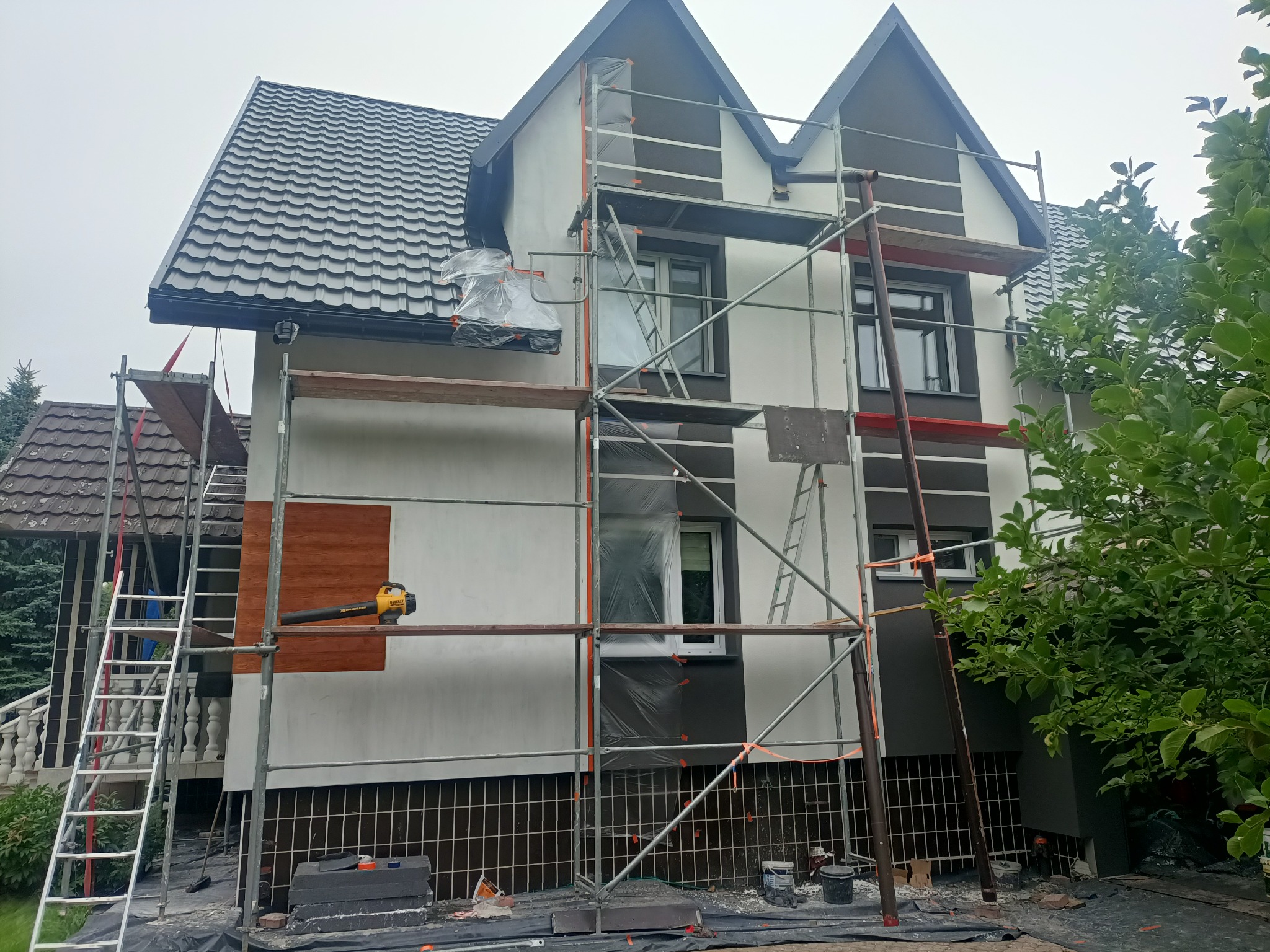 Dom w trakcie prac elewacyjnych: rusztowania, zabezpieczone okna folią, fragment elewacji z drewnopodobną okładziną, dmuchawa do liści na rusztowaniu.