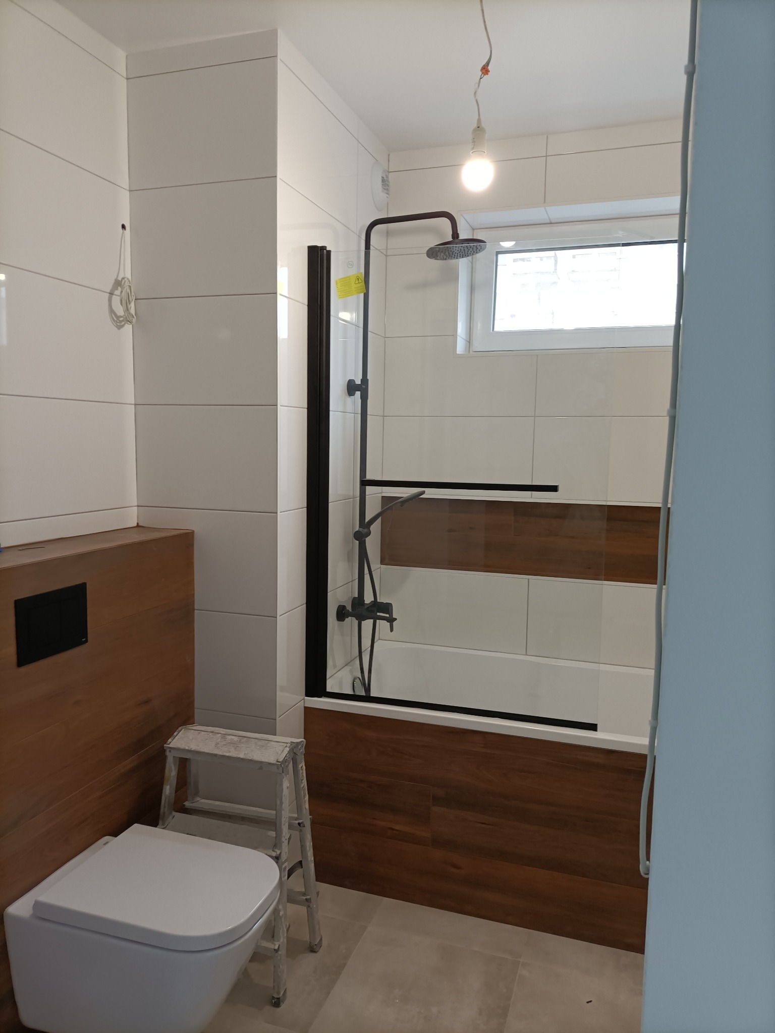 Nowoczesna łazienka w trakcie wykańczania: białe płytki, drewnopodobna zabudowa wanny i umywalki, czarna armatura, wisząca toaleta i drabinka malarska.