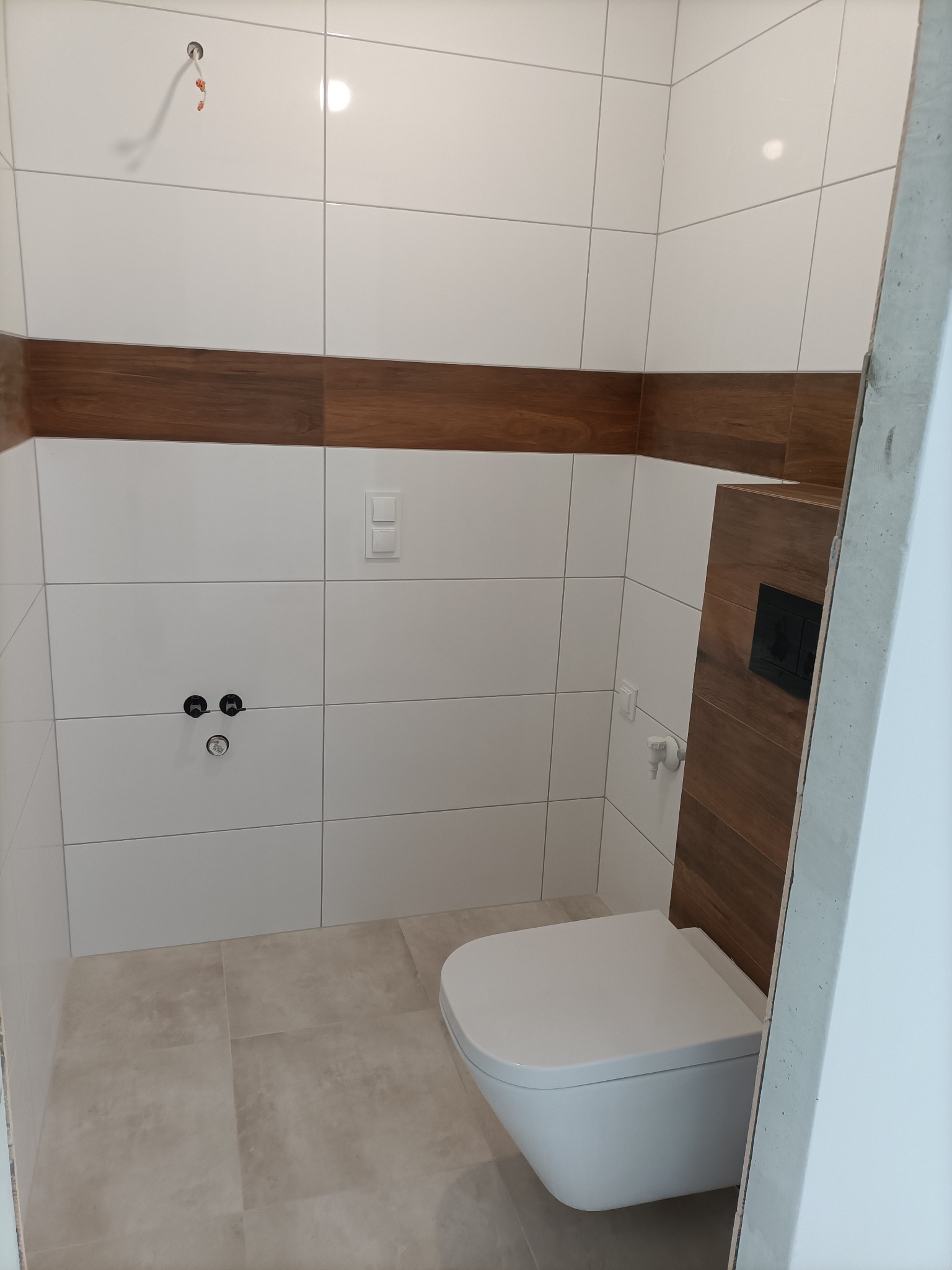 Wykończona toaleta z białymi i drewnopodobnymi płytkami, wisząca muszla klozetowa, widoczne przyłącza wodne i włącznik światła, surowy beton na krawędzi kadru.