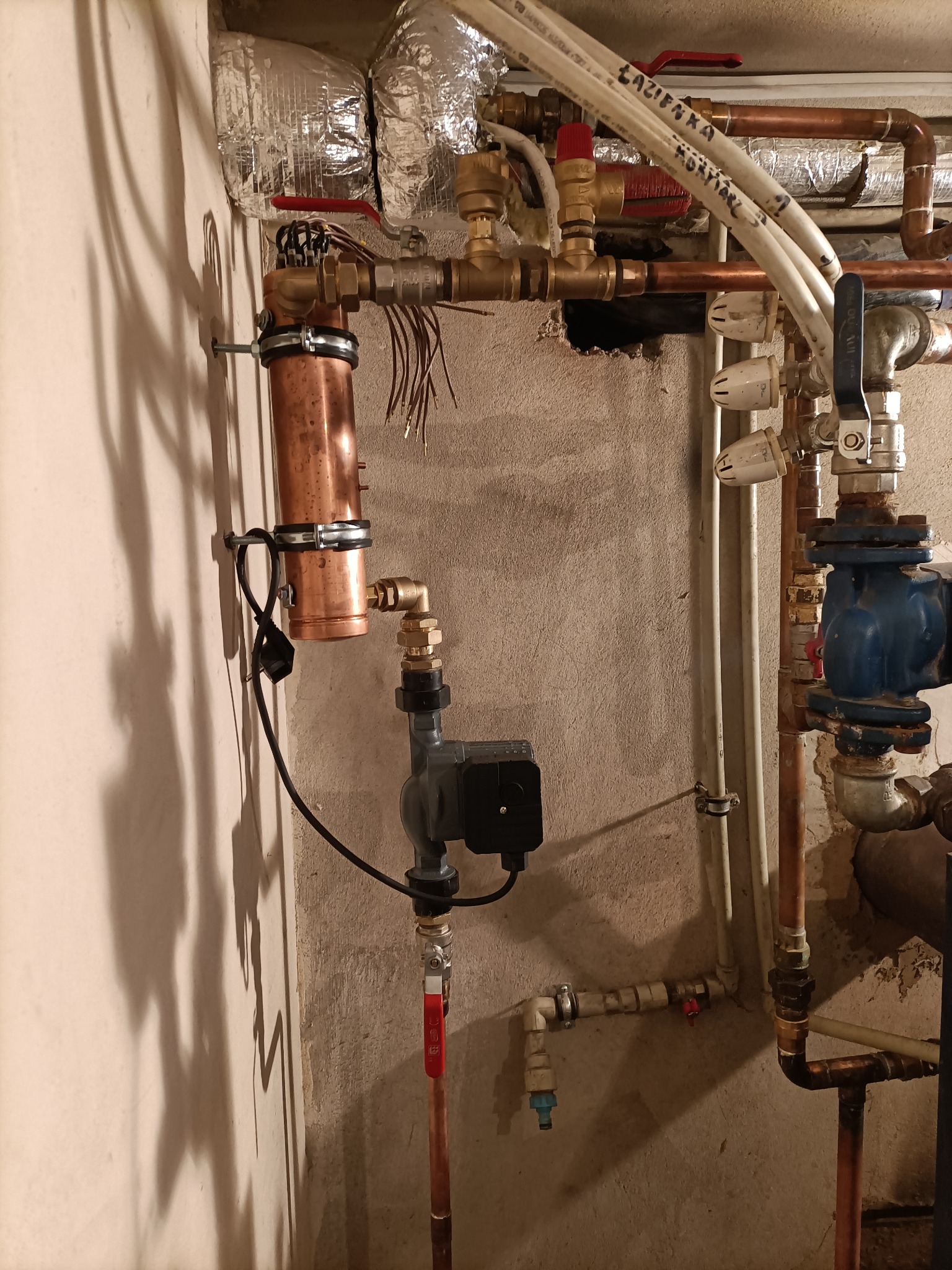 Instalacja centralnego ogrzewania z miedzianymi rurami, pompą obiegową i zaworami, widoczne przewody elektryczne i izolacja termiczna rur, ściana z widoczną fakturą.