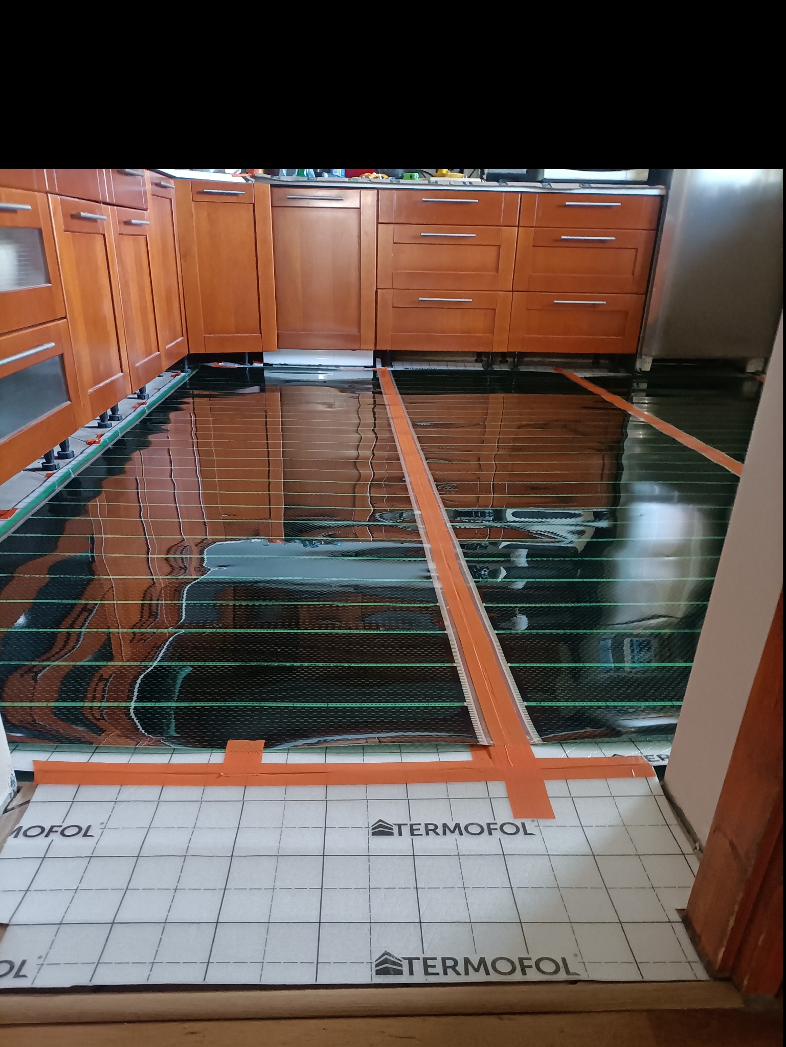 Montaż folii grzewczej Termofol na podłodze w kuchni, widoczne brązowe szafki kuchenne na regulowanych nóżkach i odblaskowa powierzchnia folii.