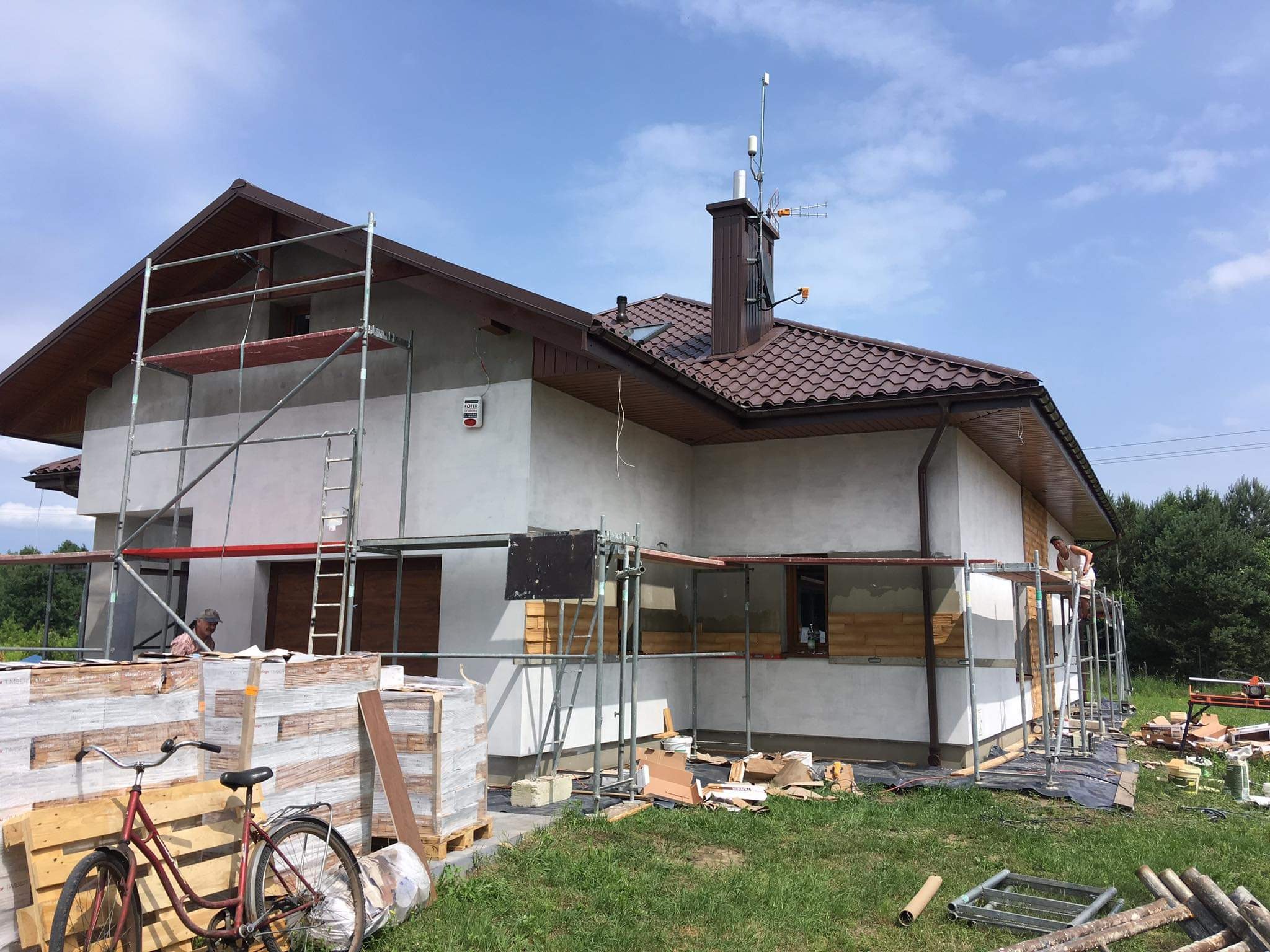 Dom jednorodzinny w trakcie prac elewacyjnych; widoczne rusztowania, ocieplenie części ścian, palety z materiałami izolacyjnymi, rower oparty o paletę, oraz pracownicy na rusztowaniu.
