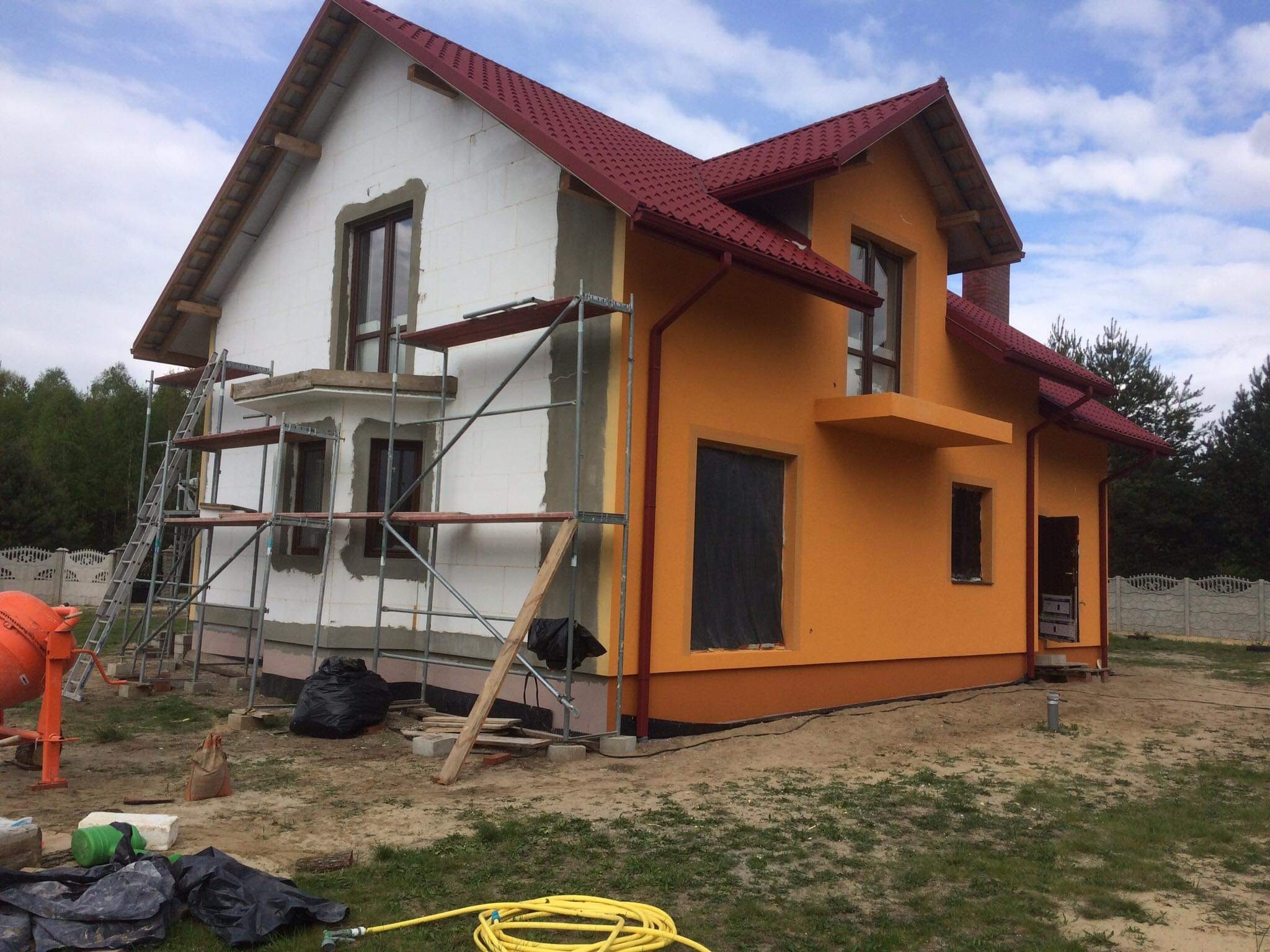 Dom jednorodzinny w trakcie prac elewacyjnych: jedna ściana ocieplona i przygotowana do malowania, druga pomalowana na pomarańczowo, widoczne rusztowania, worki z zaprawą i betoniarka.