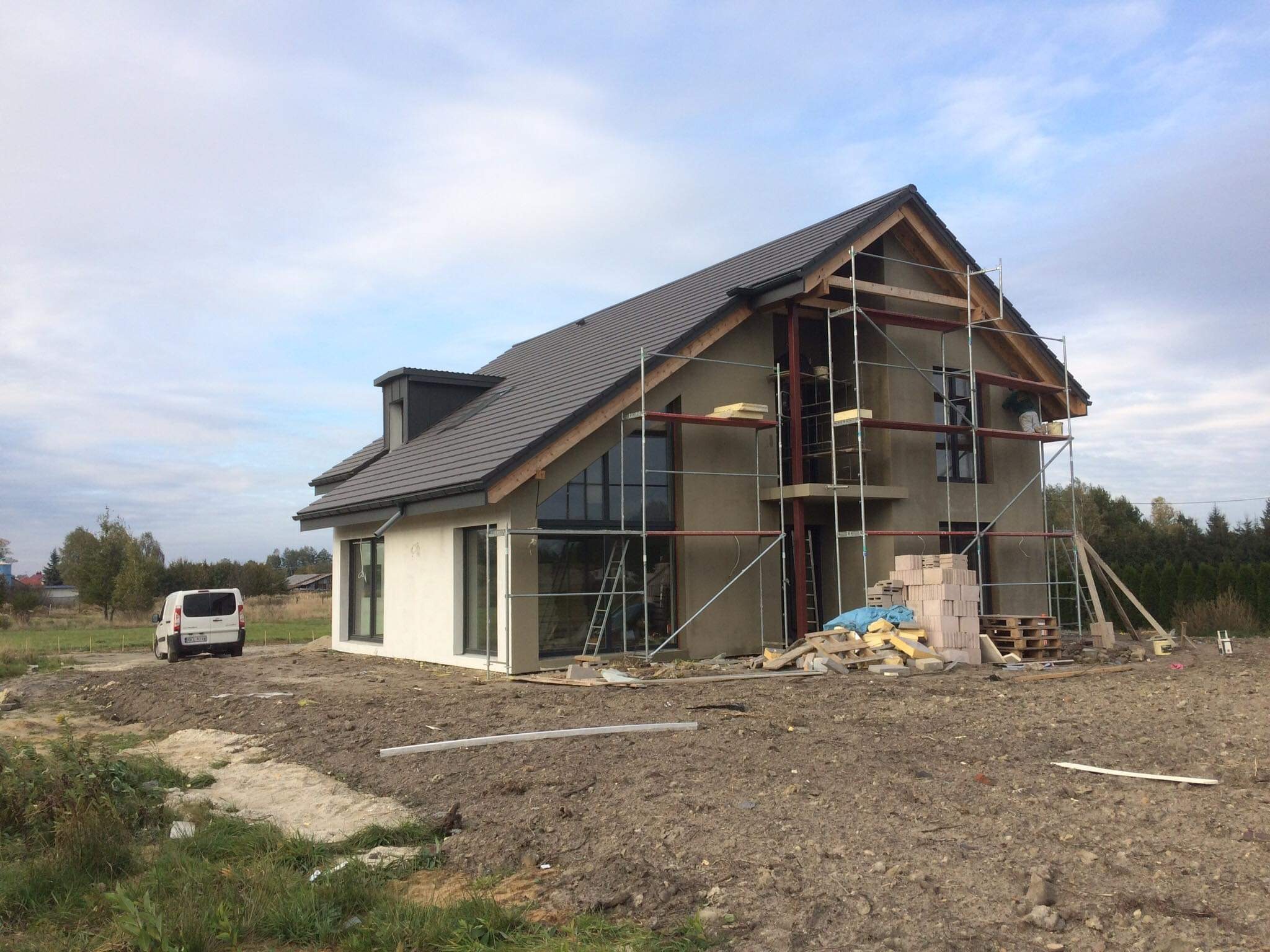 Dom jednorodzinny w trakcie ocieplania ścian zewnętrznych, widoczne rusztowania i materiały izolacyjne, na pierwszym planie niezagospodarowany teren.