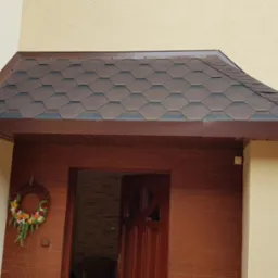 Zadaszenie nad wejściem do domu pokryte brązowym gontem bitumicznym, ściany o jasnej, piaskowej fakturze, dekoracyjny wieniec na ceglanej ścianie obok drzwi.