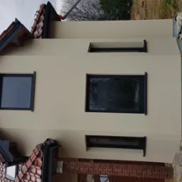 Ocieplona elewacja domu jednorodzinnego w kolorze beżowym z czarnymi oknami, widoczne fragmenty dachu z czerwonej dachówki i rury drenażowej na niezagospodarowanym terenie wokół budynku.