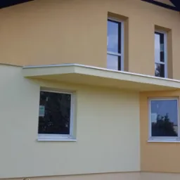 Dwukolorowa elewacja domu jednorodzinnego z widocznymi oknami i zadaszeniem nad wejściem, wykończona tynkiem strukturalnym w odcieniach beżu i kremu, z ciemnym, spadzistym dachem.