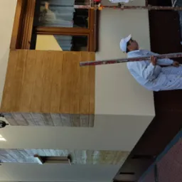 Pracownik w białym kombinezonie i czapce, stojący na rusztowaniu, mierzy poziom na elewacji budynku z drewnianymi panelami i świeżo pomalowaną, jasną ścianą.