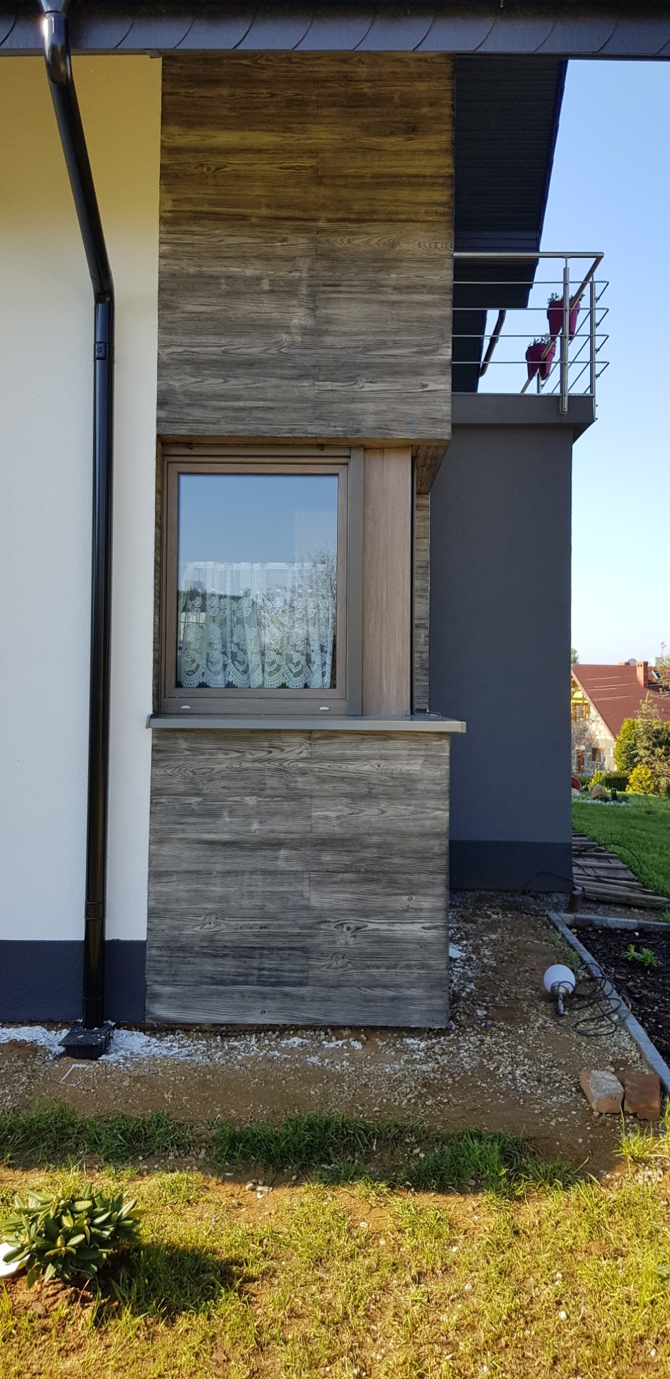 Fragment elewacji domu z oknem, wykończony panelami imitującymi stare drewno, widoczny fragment balkonu z donicami, trawnik i instalacja oświetleniowa w gruncie.
