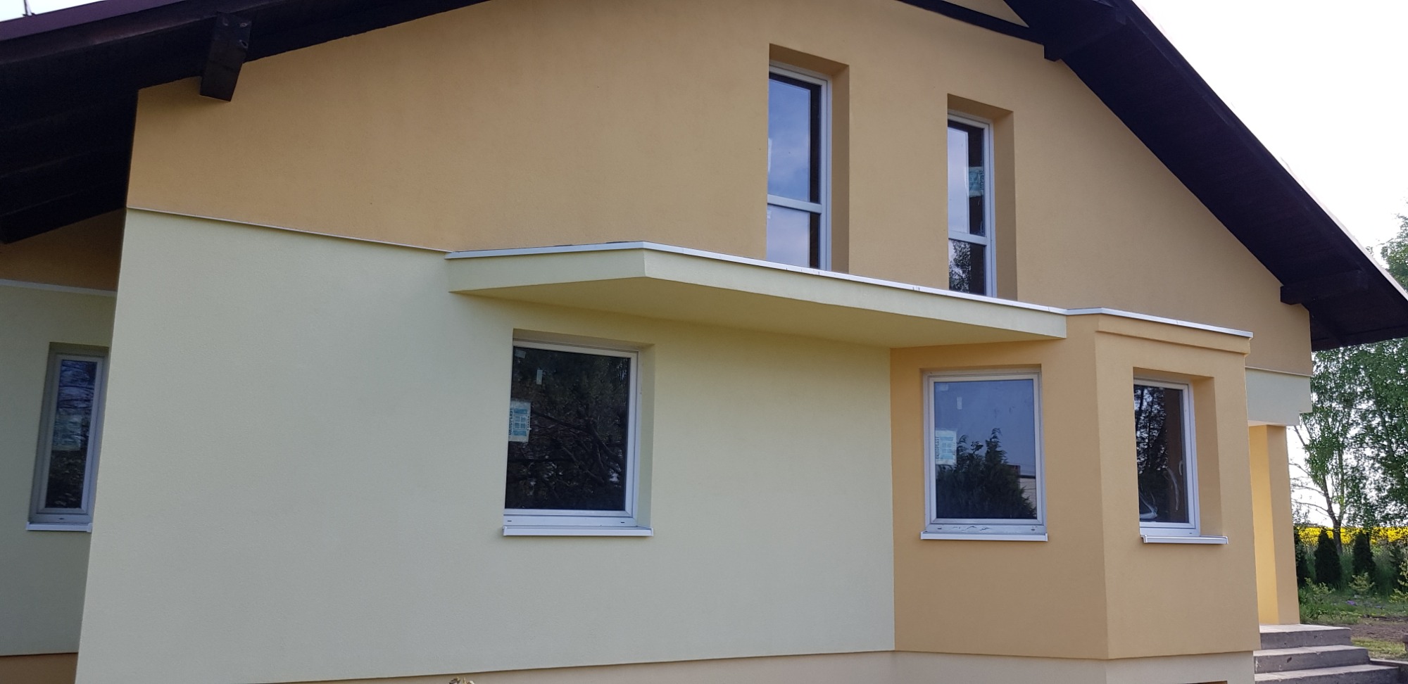 Dwukolorowa elewacja domu jednorodzinnego z widocznymi oknami i zadaszeniem nad wejściem, wykończona tynkiem strukturalnym w odcieniach beżu i kremu, z ciemnym, spadzistym dachem.