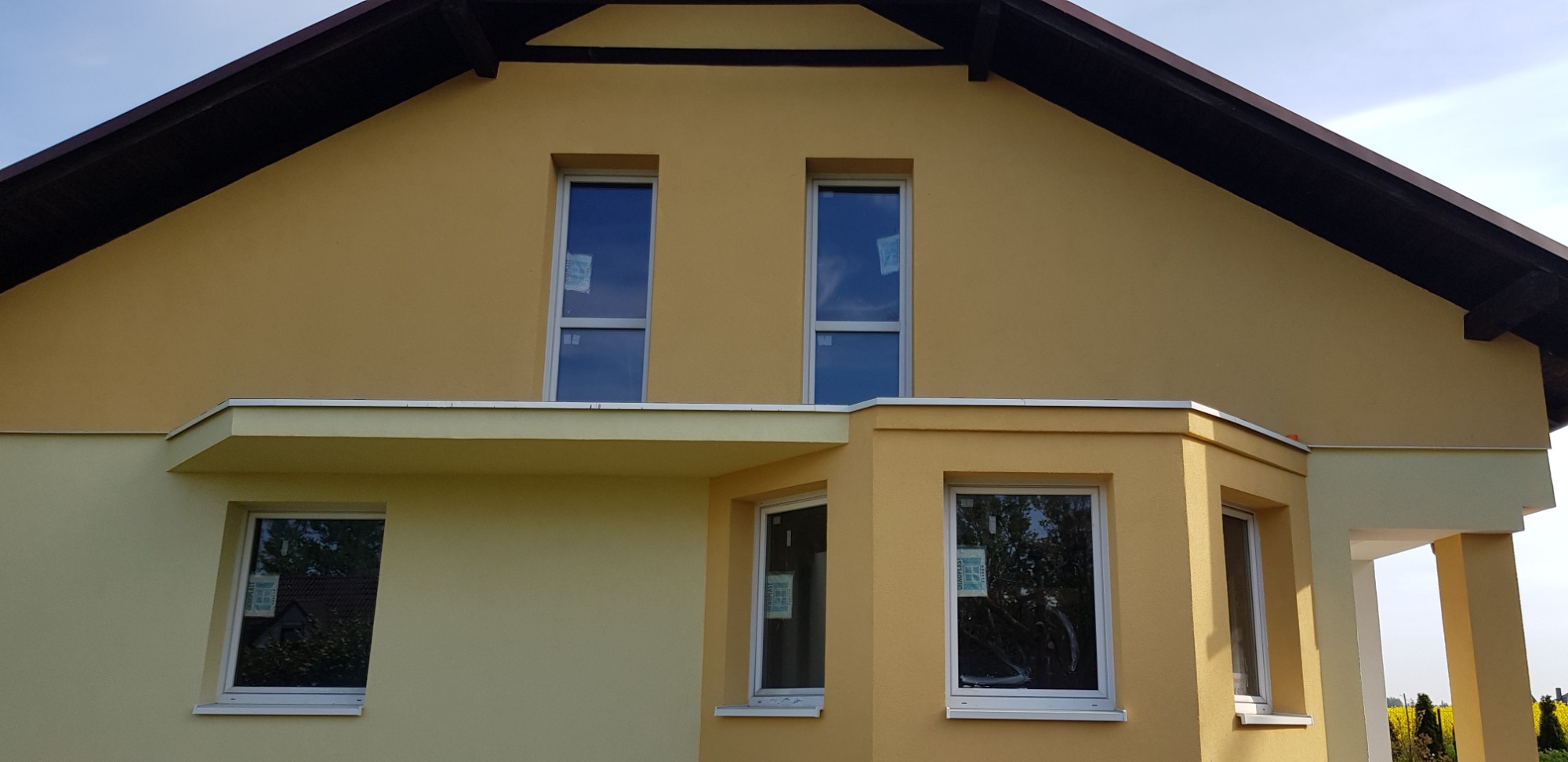 Żółta elewacja domu jednorodzinnego z nowymi oknami, widoczne naklejki na szybach, jasna podbitka dachowa i fragment kolumn werandy.