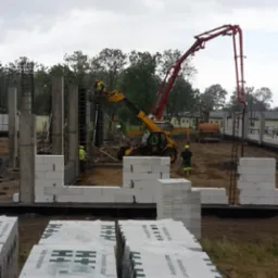 Budowa domu z betonu komórkowego: ściany parteru w trakcie wznoszenia, zbrojenia i betonowe słupy, pompa do betonu w akcji, palety z bloczkami na pierwszym planie.