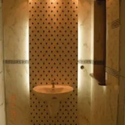 Umywalka na postumencie z baterią, na tle ściany z mozaiką w łazience oświetlonej punktowo, z marmurowymi płytkami i dekoracyjnym pasem na podłodze.