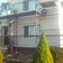 Prace elewacyjne w Żninie: robotnicy na rusztowaniu tynkują jasną ścianę domu z oknami. Widoczny styropian i wiadro z zaprawą.