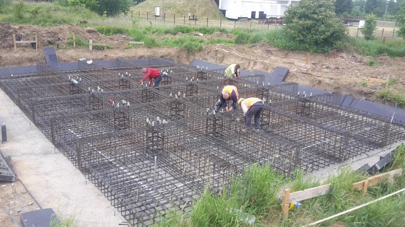 Ekipa montuje zbrojenie fundamentów pod budynek na działce. Widoczne stalowe pręty, szalunki i pracownicy w odblaskowych kamizelkach. Prace ziemne w tle.