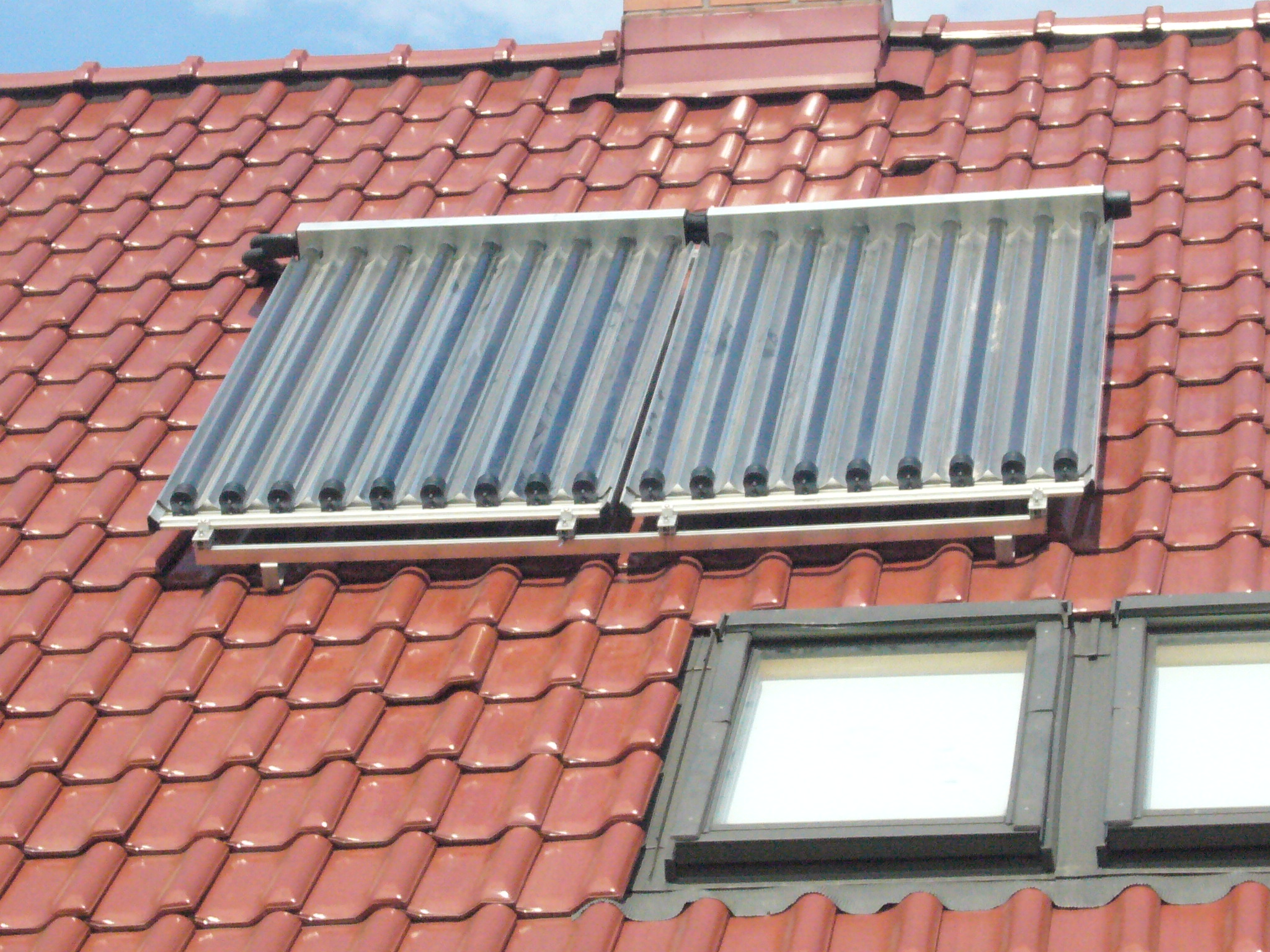 Dwa panele kolektorów słonecznych zamontowane na dachu z czerwonej dachówki, obok okna dachowego.