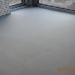 Świeżo wylana, gładka, szara posadzka jastrychowa w rogu pomieszczenia z oknem, widoczna folia izolacyjna przy ścianie.