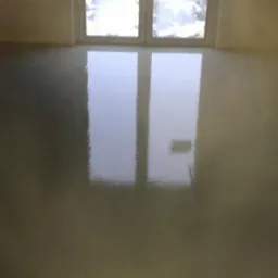 Świeżo wylana, błyszcząca posadzka w jasnym pomieszczeniu z oknem, odbijająca światło, widoczne delikatne nierówności powierzchni.