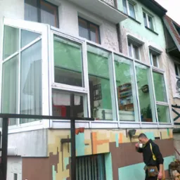 Przeszklona weranda na piętrze budynku z widocznymi zasłonami, regałem z książkami i roślinami doniczkowymi wewnątrz, z mężczyzną pijącym kawę na zewnątrz i stosem drewna obok.