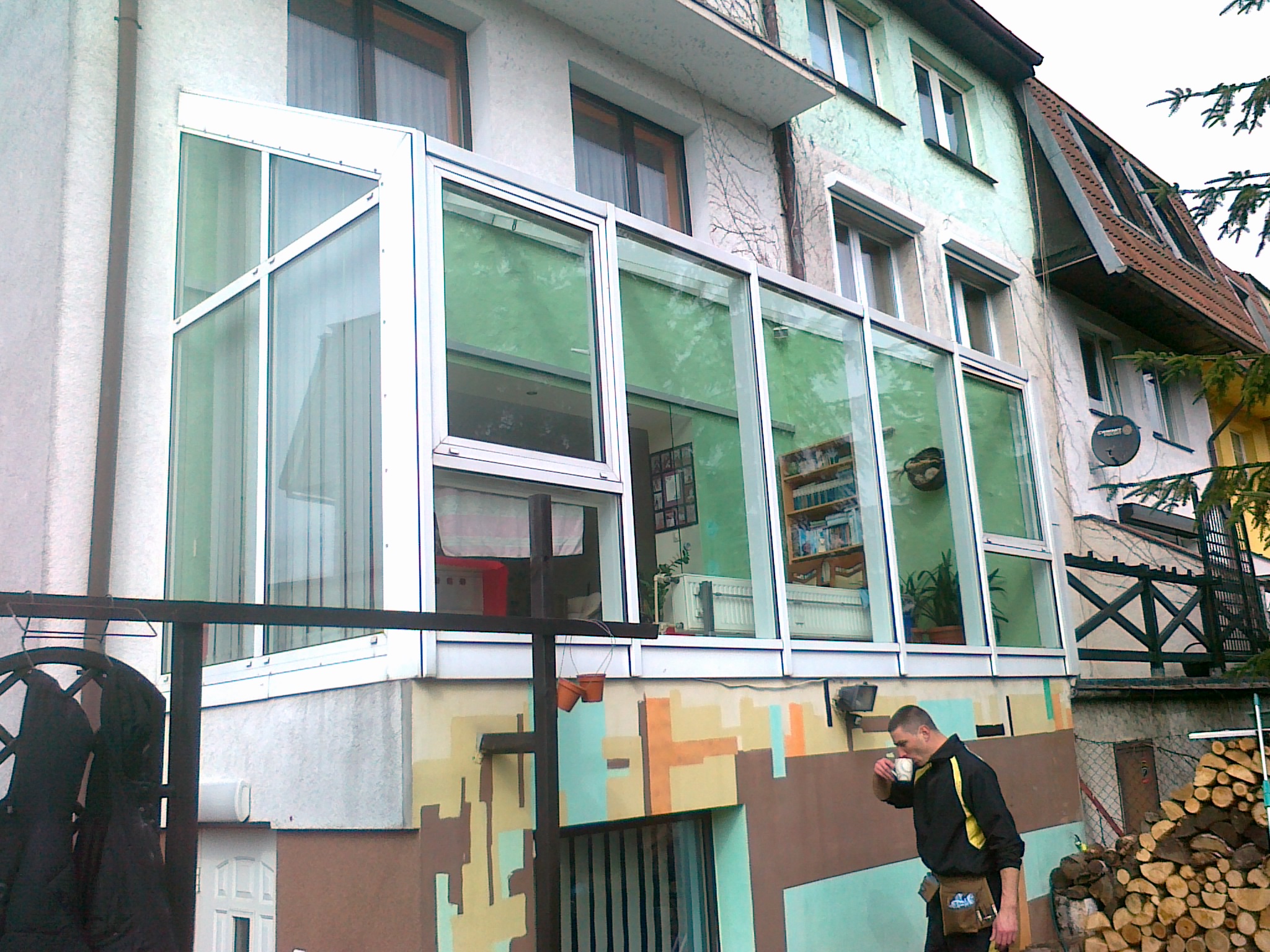 Przeszklona weranda na piętrze budynku z widocznymi zasłonami, regałem z książkami i roślinami doniczkowymi wewnątrz, z mężczyzną pijącym kawę na zewnątrz i stosem drewna obok.